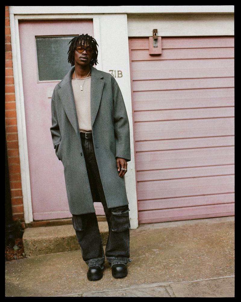 Man in gray coat poses on sidewalk in front of pink door with house number visible, dressed in modern urban fashion.