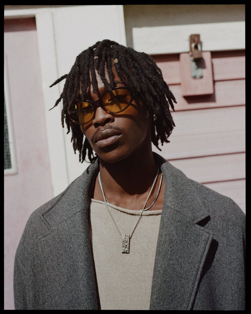 Portrait of an African American man with dreadlocks wearing translucent yellow glasses, a grey coat and standing outdoors.