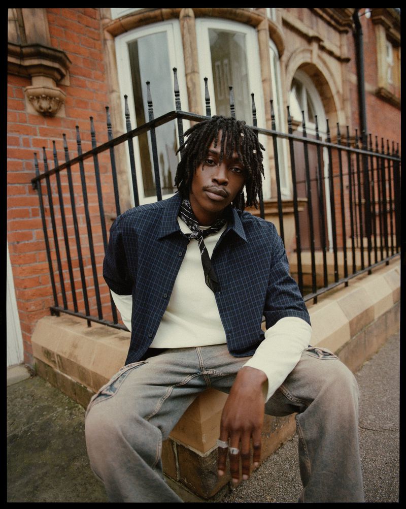 A young adult male in a casual fashion outfit sits on a ledge in front of a brick building with iron fencing, captured in a medium shot.