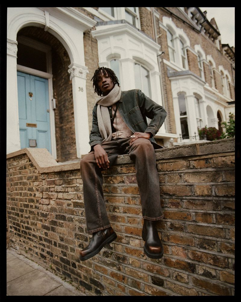 A stylish adult male sits on a brick ledge in front of a town house wearing a dark jacket, tan scarf, and dark pants with brown boots.