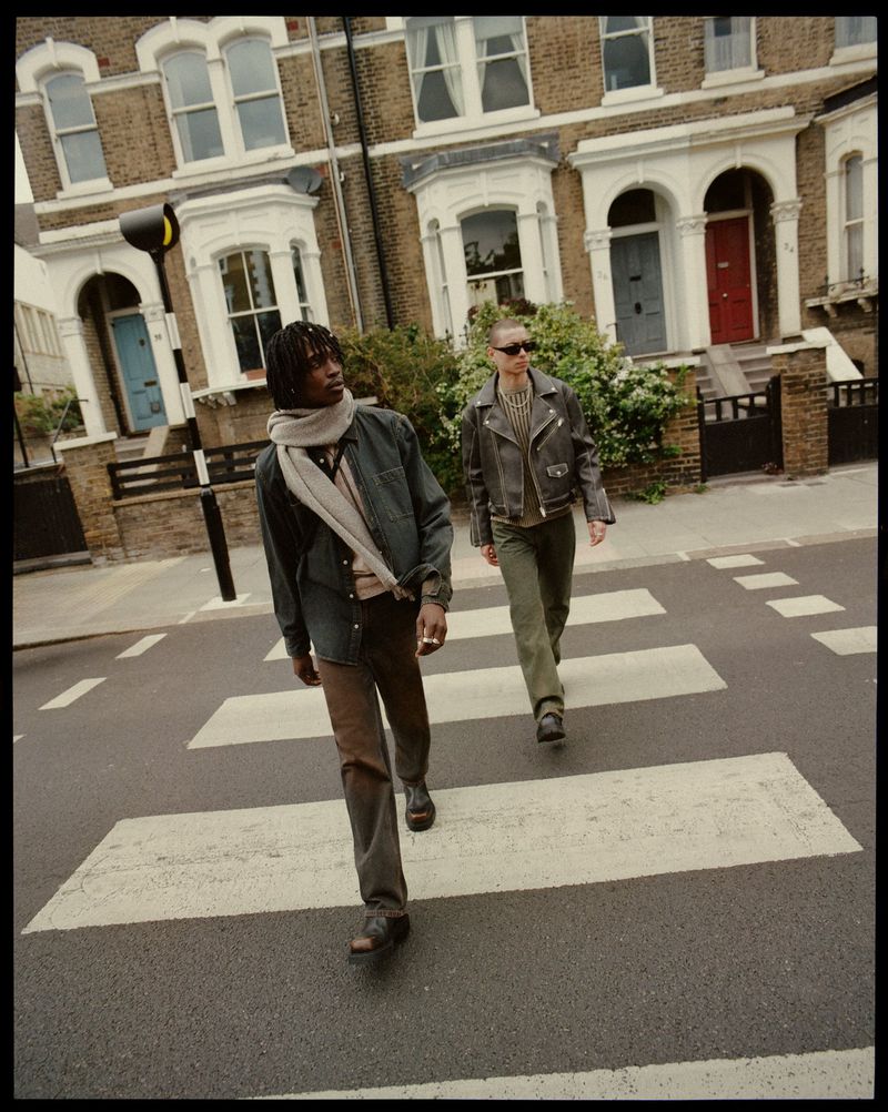 Two adult men walking across a crosswalk wearing casual clothing, including a jacket, jeans, and scarves, with buildings in the background.