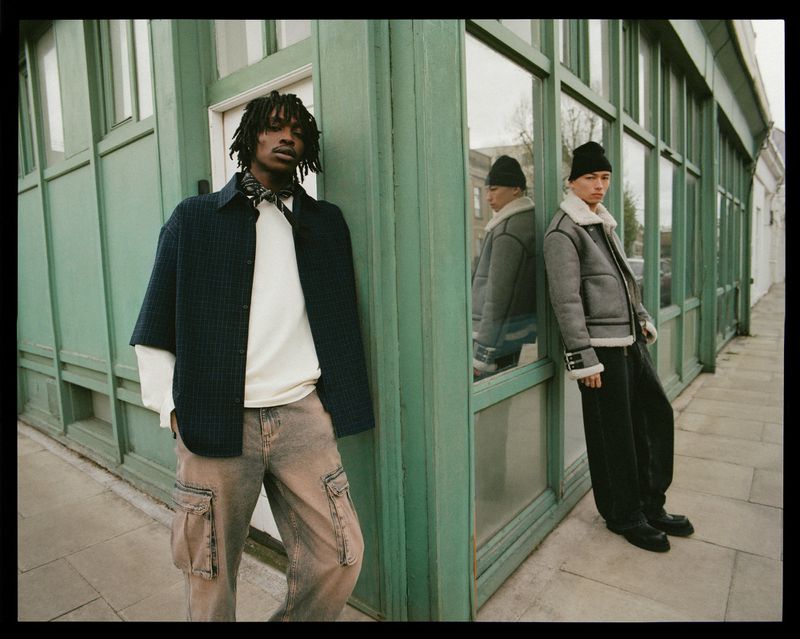 Two young men in casual fashion stand outdoors against a green building. One wears a navy shirt jacket and cargo pants; the other a gray jacket.