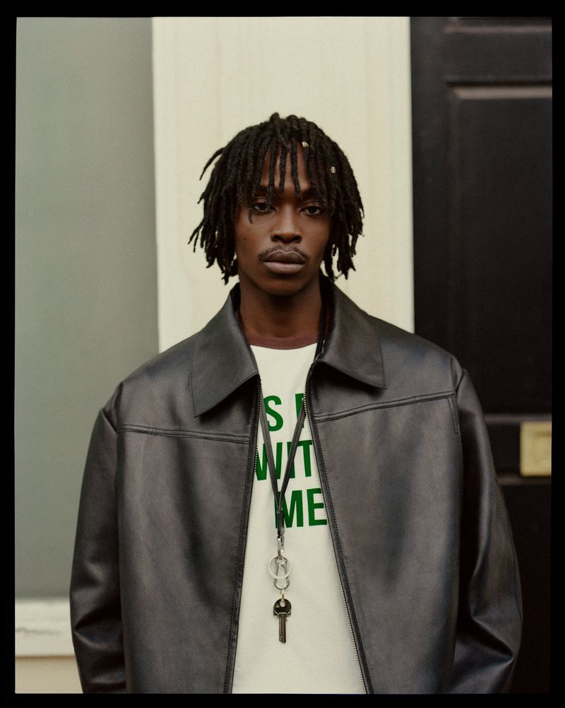 Portrait of a black male model with dreadlocks wearing a black leather jacket and a white t-shirt against a neutral background.