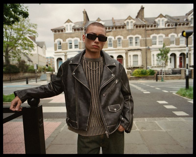 Man in leather jacket and sunglasses stands in urban street setting. Buildings in background. Fashion editorial with modern style.