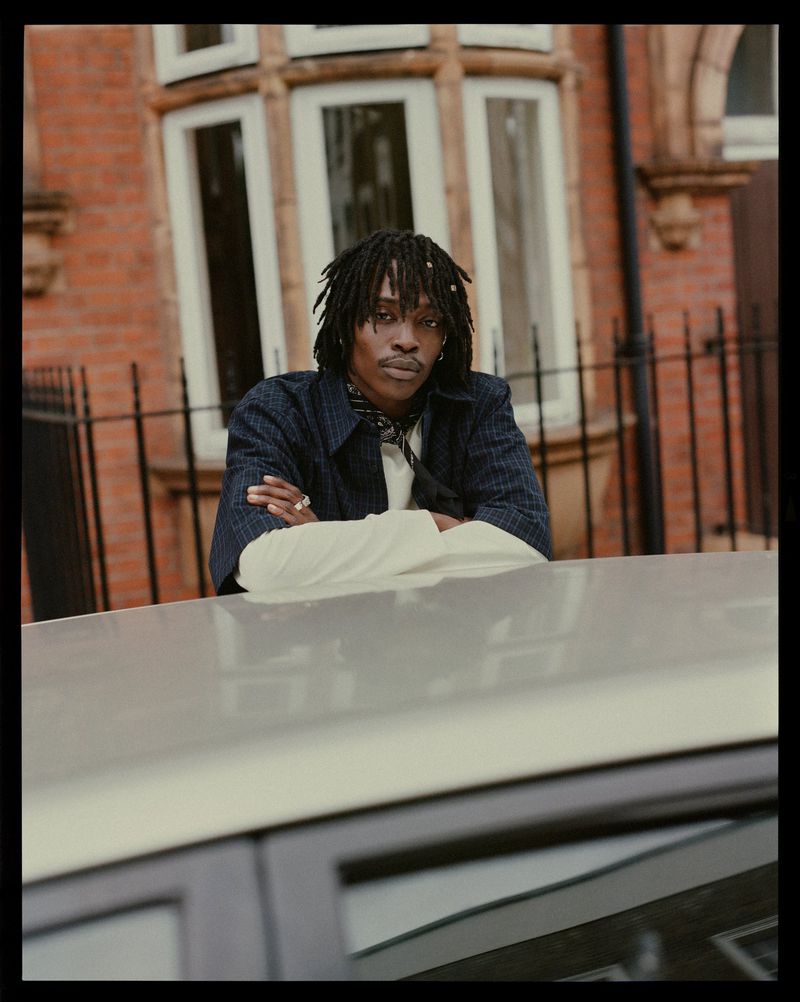 Portrait of a black man with dreadlocks sitting on the hood of a car in front of a red brick building in an urban setting, fashionable and stylish.