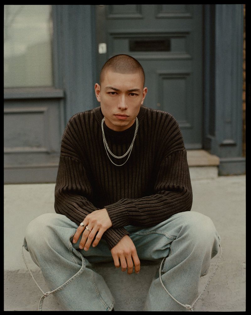 Portrait of a young adult man sitting on steps outdoors, wearing a dark brown knit sweater, light blue jeans, and silver necklaces.