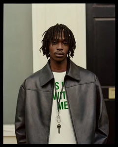 Male model wearing an open black leather jacket over a graphic white t-shirt and key lanyard outdoors by a black door.
