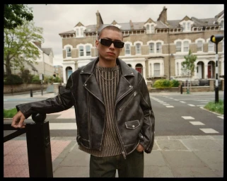 A young man wearing a black leather jacket and sunglasses leans against a railing on a London street.