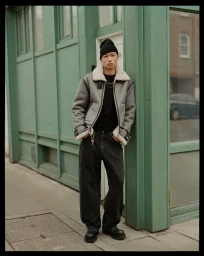 Model Nicolas Yoshi wearing a grey shearling jacket, black beanie, and wide-leg jeans standing beside a green storefront.