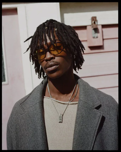 Black man with dreadlocks wearing yellow-tinted sunglasses, a grey overcoat, silver pendant necklace, and cream knit top.