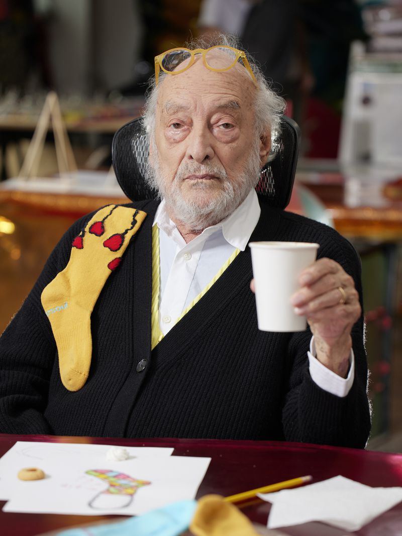 Elderly man seated indoors with glasses on his head holding a cup, wearing a cardigan with a sock over his shoulder.