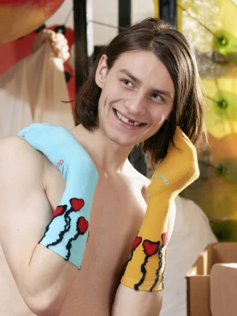 Shirtless person smiles wearing blue and yellow sock-sleeves with black vines and red heart designs against a blurred background.