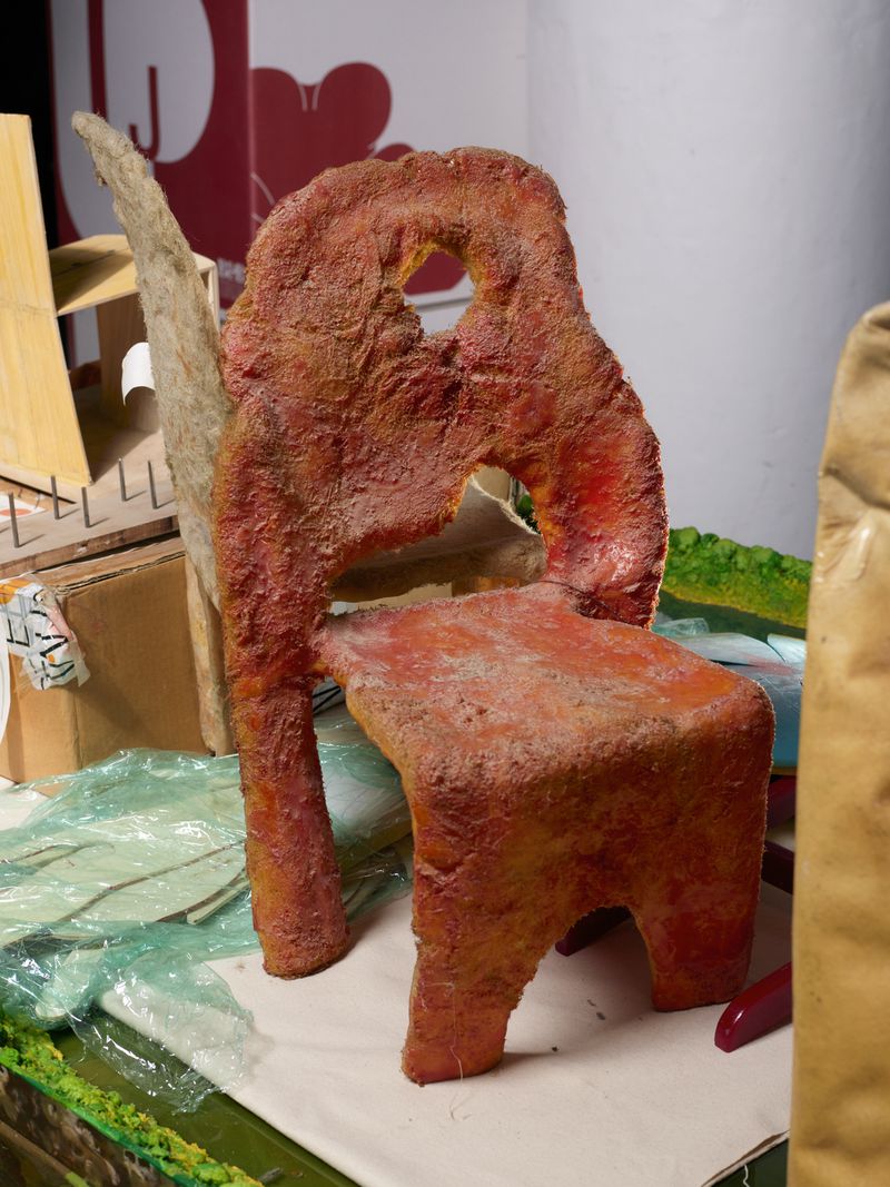 Artisanal chair sculpture with an irregular, textured surface in shades of red and brown, featuring carved-out sections and a rustic aesthetic.