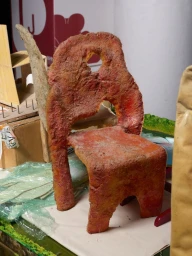 Textured reddish-orange sculptural miniature chair surrounded by workshop materials and model pieces.