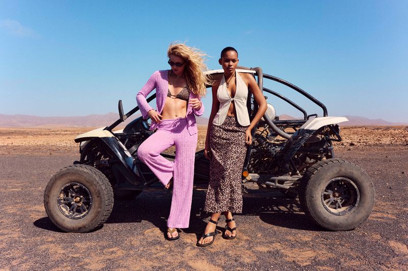 Two fashion models pose with a recreational buggy in the desert against a blue sky, showcasing stylish summer outfits and accessories.
