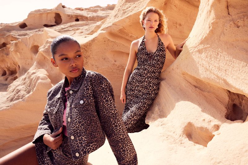 Two fashion models pose in animal print clothing in a desert landscape with beige rock formations, captured in outdoor fashion photography.