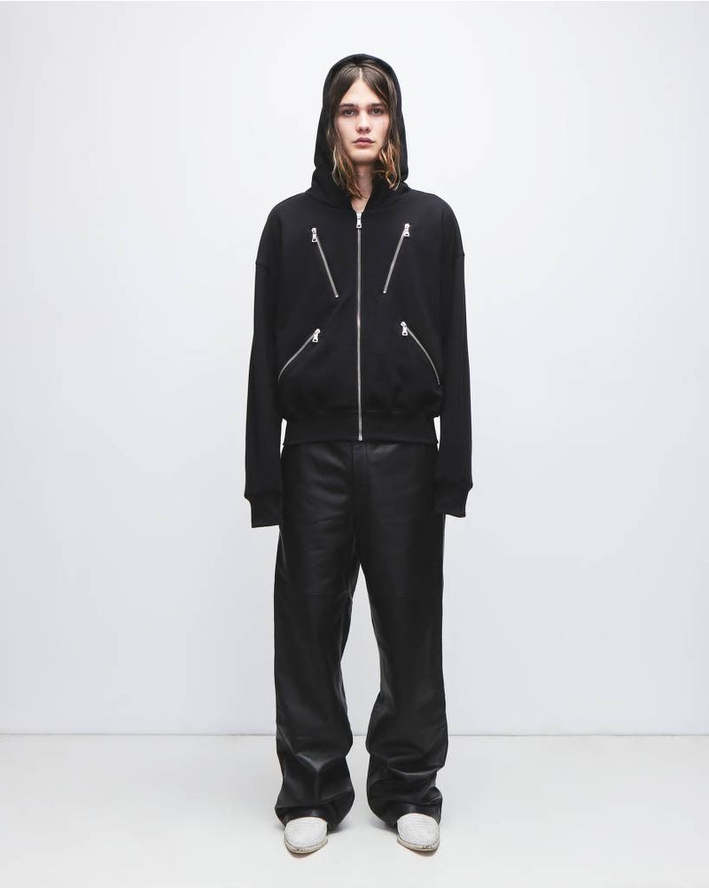 Full-length shot of an adult male model in a black zip-up hooded jacket with metal zippers, black pants, and white sneakers in a photo studio.
