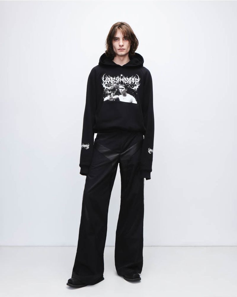 Person in black hoodie and wide-leg pants poses against a bright background, showcasing fashion apparel in a studio setting.