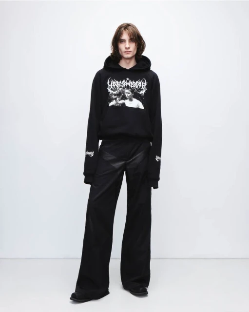 Person in black hoodie and wide-leg pants poses against a bright background, showcasing fashion apparel in a studio setting.