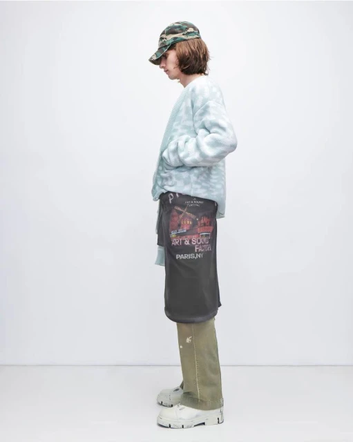 A person is standing indoors wearing a camouflage hat, light blue sweater, olive pants, and thick sole shoes against a white background.