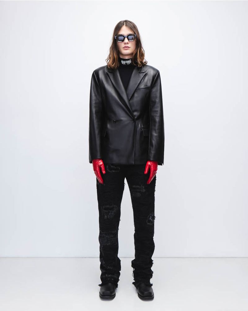 Adult male fashion model in a black leather jacket, black ripped jeans, red gloves, and sunglasses poses in a photo studio.
