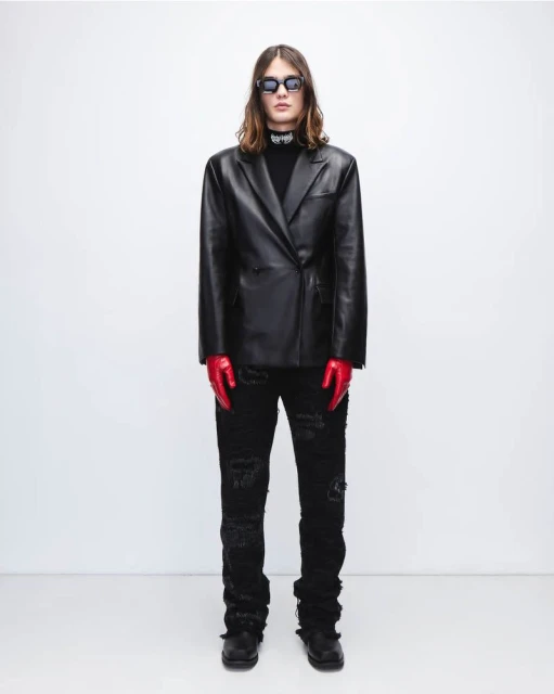 Adult male fashion model in a black leather jacket, black ripped jeans, red gloves, and sunglasses poses in a photo studio.