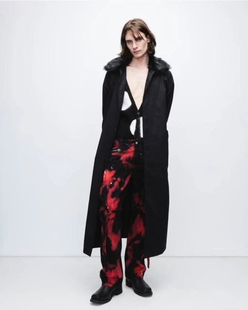 Male fashion model in black coat and red tie-dye pants posing in a photo studio with a white backdrop and wearing black leather boots.