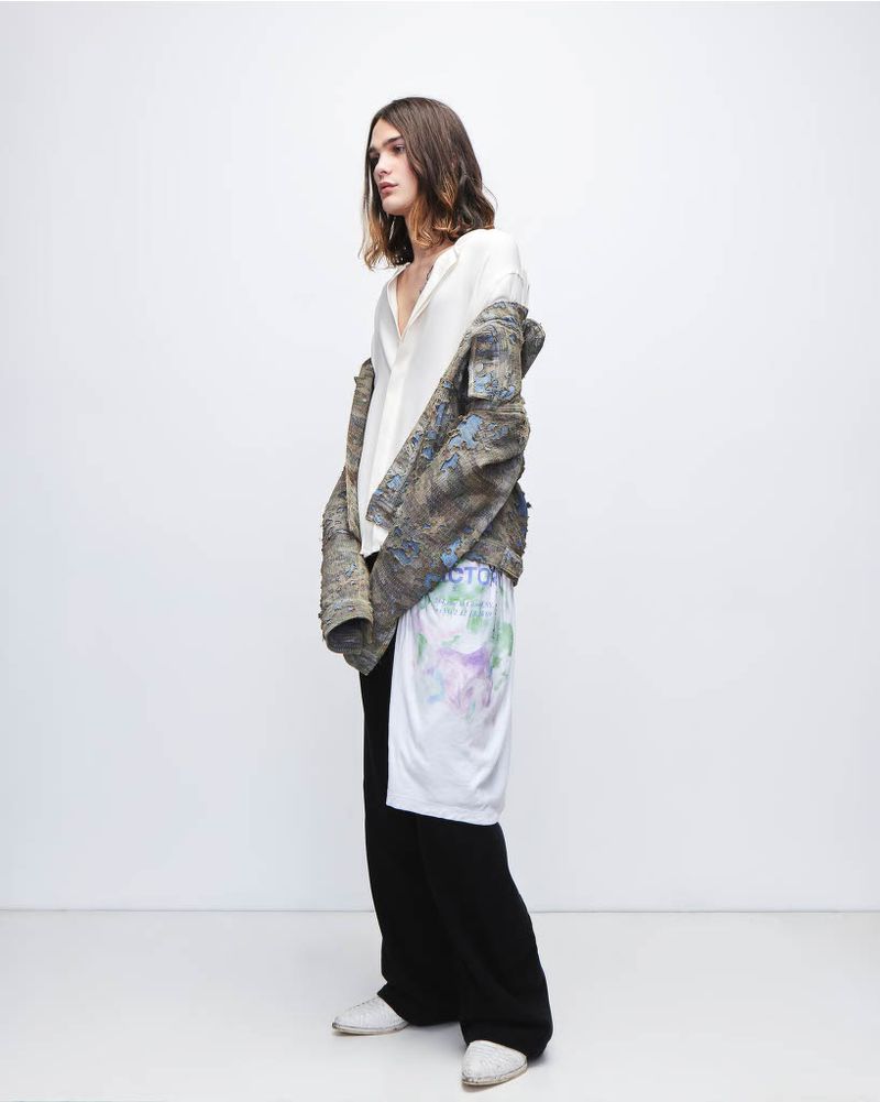 A slender person in a white shirt, jacket, and black pants stands in a photo studio with a white backdrop, looking off to the side.