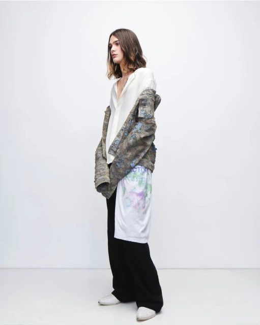 A slender person in a white shirt, jacket, and black pants stands in a photo studio with a white backdrop, looking off to the side.