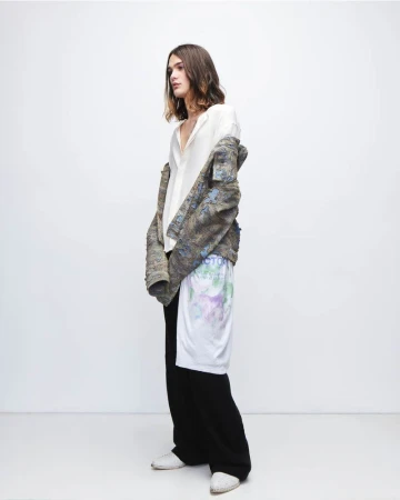 A slender person in a white shirt, jacket, and black pants stands in a photo studio with a white backdrop, looking off to the side.