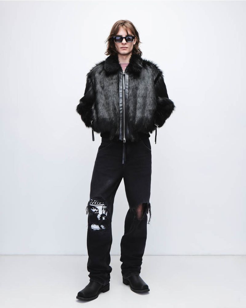 Full-length shot of a man in a black fur jacket, distressed jeans, boots, and sunglasses against a white background in a studio setting.
