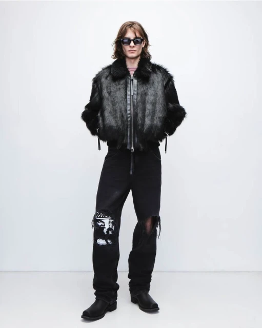 Full-length shot of a man in a black fur jacket, distressed jeans, boots, and sunglasses against a white background in a studio setting.