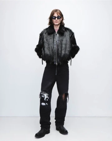 Full-length shot of a man in a black fur jacket, distressed jeans, boots, and sunglasses against a white background in a studio setting.
