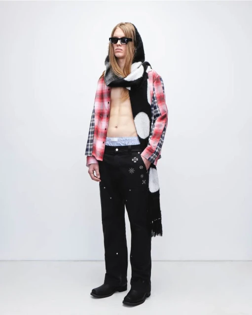 An adult male poses in a photo studio wearing sunglasses, a plaid shirt, patterned scarf, and embellished pants against a white backdrop.