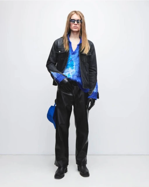 Full-length shot of a person with long blonde hair wearing a tie-dye shirt, jacket, leather pants, and sunglasses against a white backdrop.