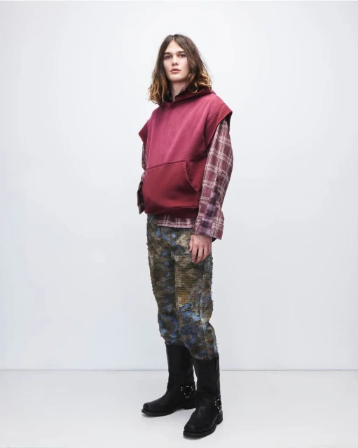 Full shot of a male fashion model wearing a sleeveless hoodie, plaid shirt, patterned pants, and black boots in a studio setting.