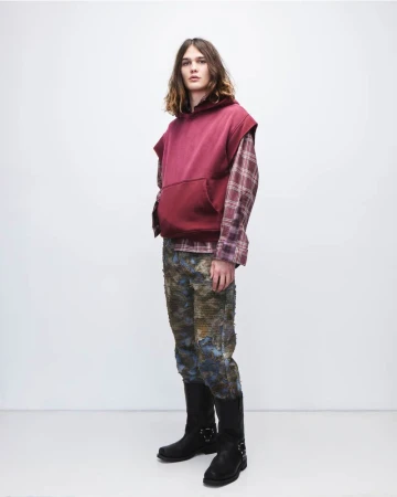 Full shot of a male fashion model wearing a sleeveless hoodie, plaid shirt, patterned pants, and black boots in a studio setting.