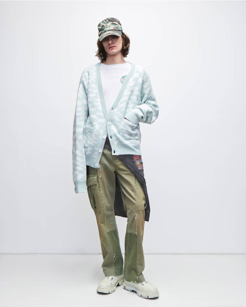 Man in patterned cardigan, t-shirt, cargo pants, baseball cap, and platform shoes poses in a studio with a neutral background.