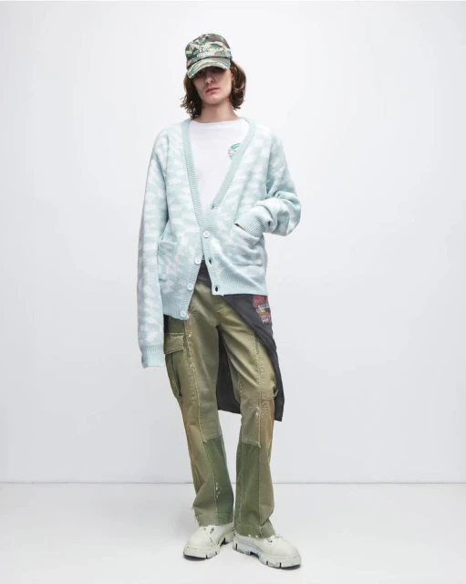 Man in patterned cardigan, t-shirt, cargo pants, baseball cap, and platform shoes poses in a studio with a neutral background.