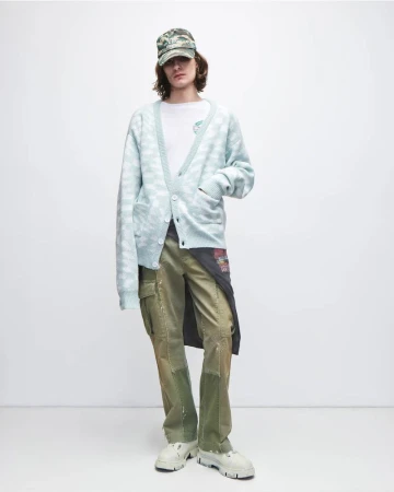 Man in patterned cardigan, t-shirt, cargo pants, baseball cap, and platform shoes poses in a studio with a neutral background.