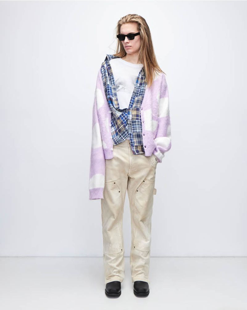 A young adult poses in a relaxed studio shot wearing a cardigan, open plaid shirt, t-shirt, pants, chunky shoes and dark sunglasses.