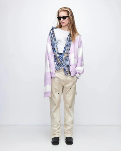 A young adult poses in a relaxed studio shot wearing a cardigan, open plaid shirt, t-shirt, pants, chunky shoes and dark sunglasses.