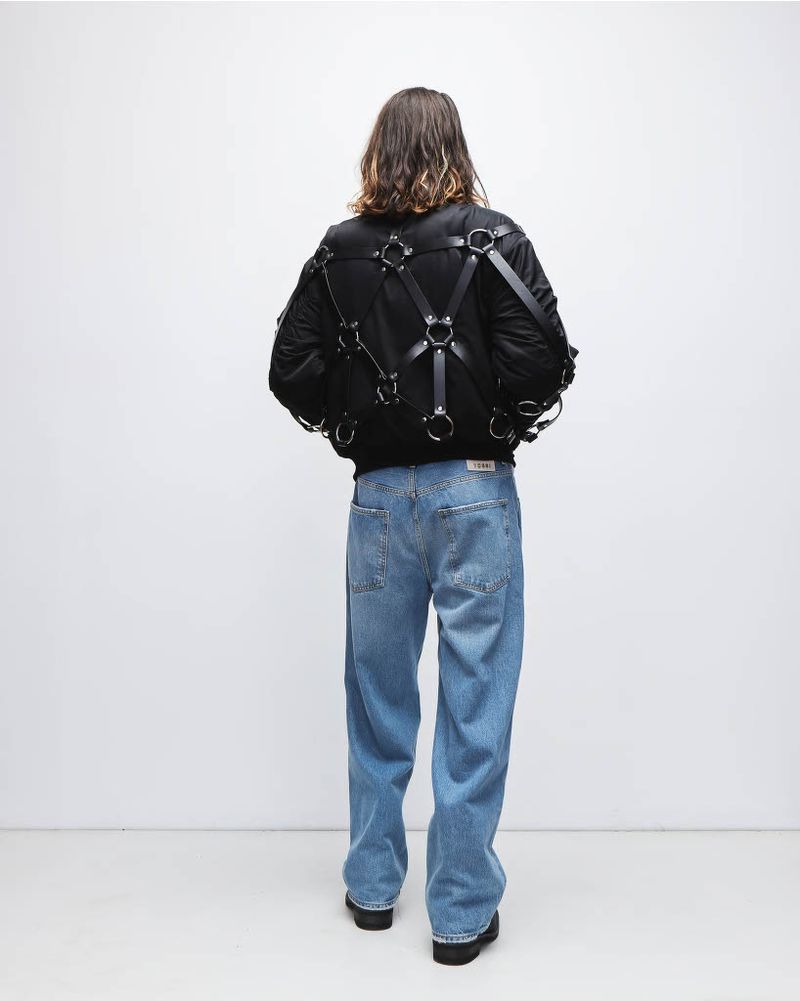 Person wearing a black jacket with harness detail and blue jeans stands against a white backdrop in a studio setting.