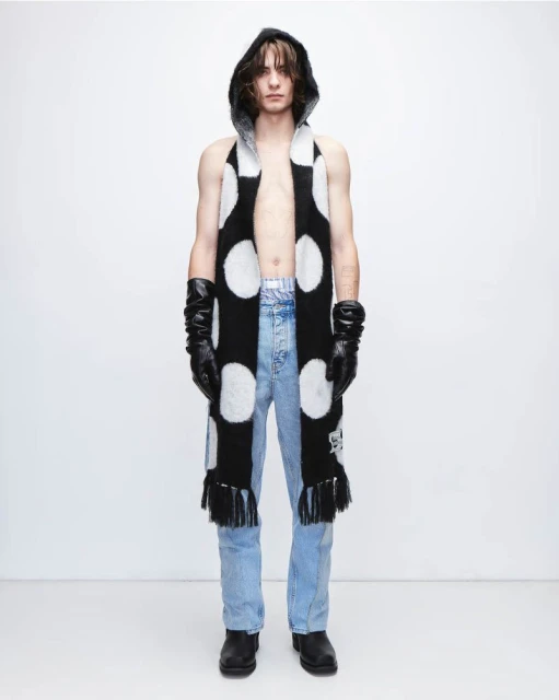 A person wearing a black and white polka dot scarf, gloves, jeans, and boots stands in front of a white backdrop in a studio shot.