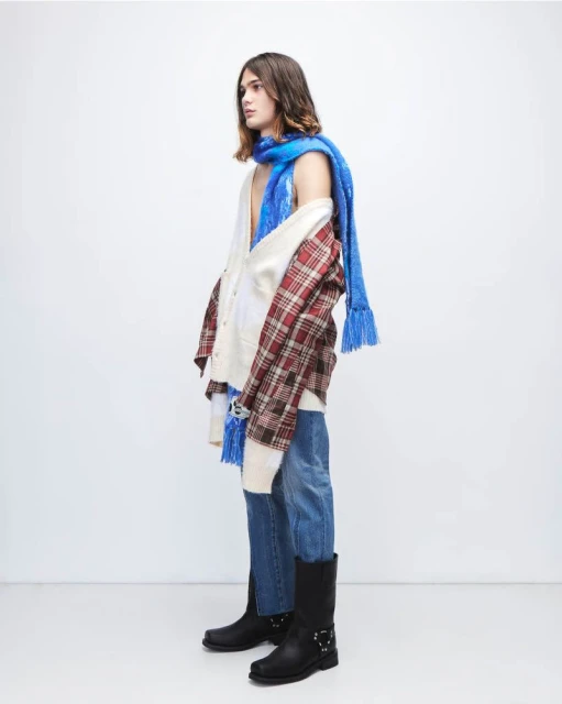 Side view of a young adult male wearing a blue scarf, patterned shirt, white cardigan, blue jeans, and black boots in a bright studio.