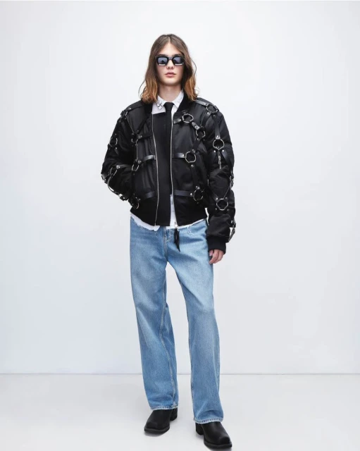 Full-length shot of a young adult in a black bomber jacket, jeans, and black boots against a bright white background. Fashion editorial.