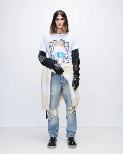A slender male fashion model poses against a white backdrop, showcasing a casual graphic tee, distressed denim, and black leather gloves.