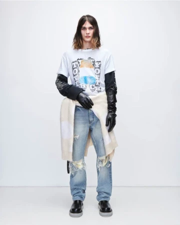 A slender male fashion model poses against a white backdrop, showcasing a casual graphic tee, distressed denim, and black leather gloves.