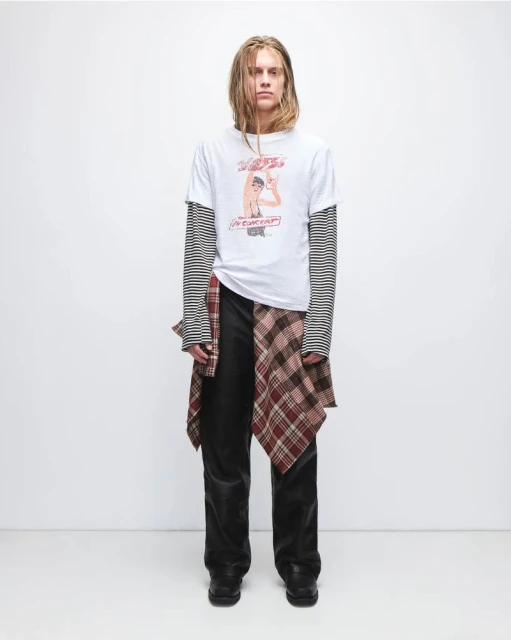 Full-length portrait of a slender adult man wearing a graphic tee, long-sleeve top, dark denim jeans, and shoes in a photo studio.