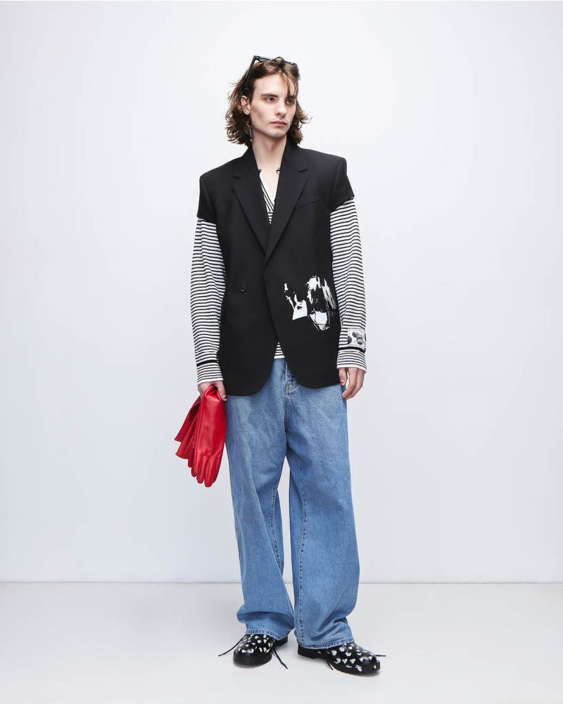 Full-length shot of a young adult male fashion model in an oversized black blazer, jeans, and black shoes against a white backdrop.
